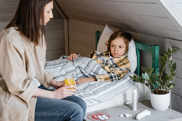 Child receiving medicine from his mother; sick child lying in bed with medication on table