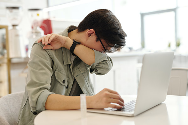 Asian man with allergies sneezing into the crook of his elbow while working in his home office