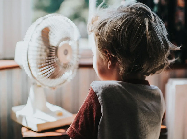 Child standing in front of a fan indoors to cool off during summer