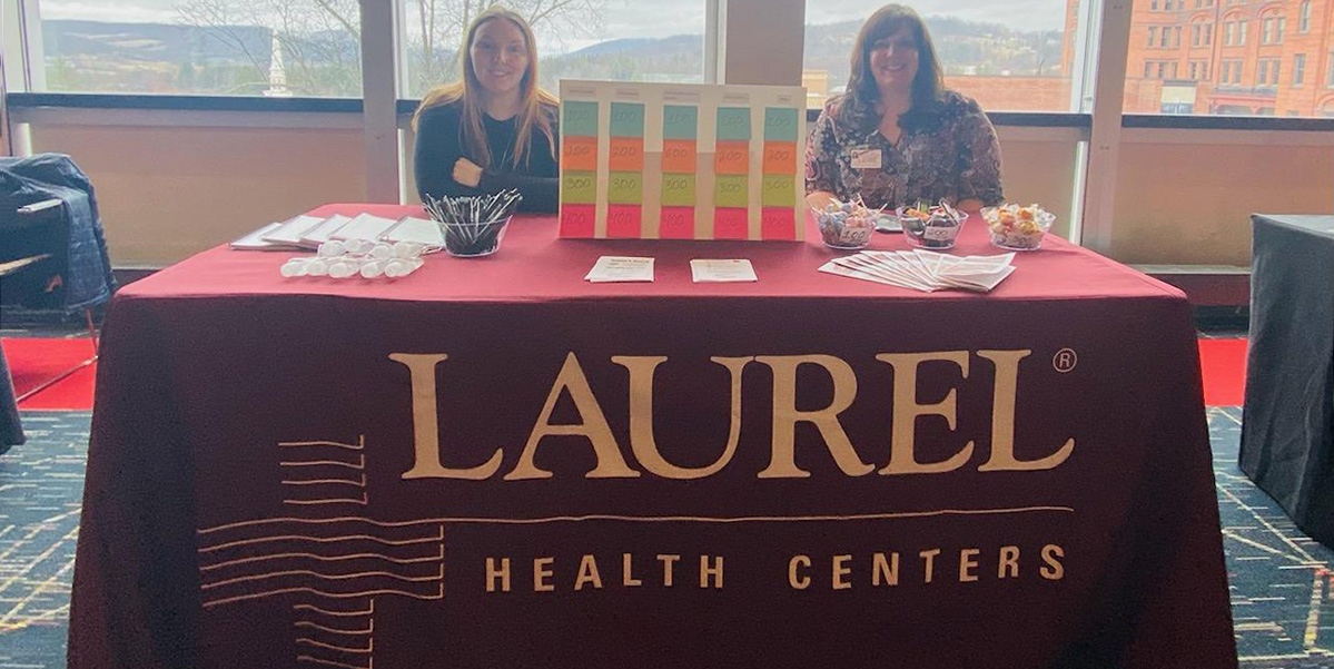 Julia York, Quality Manager Nurse, and Amy Bowens, Director of Operations, seated at Laurel Health's booth at the Women's Health & Resources Fair at Mansfield University with an LHC burgundy tablecloth, giveaways, pamphlets, and prizes