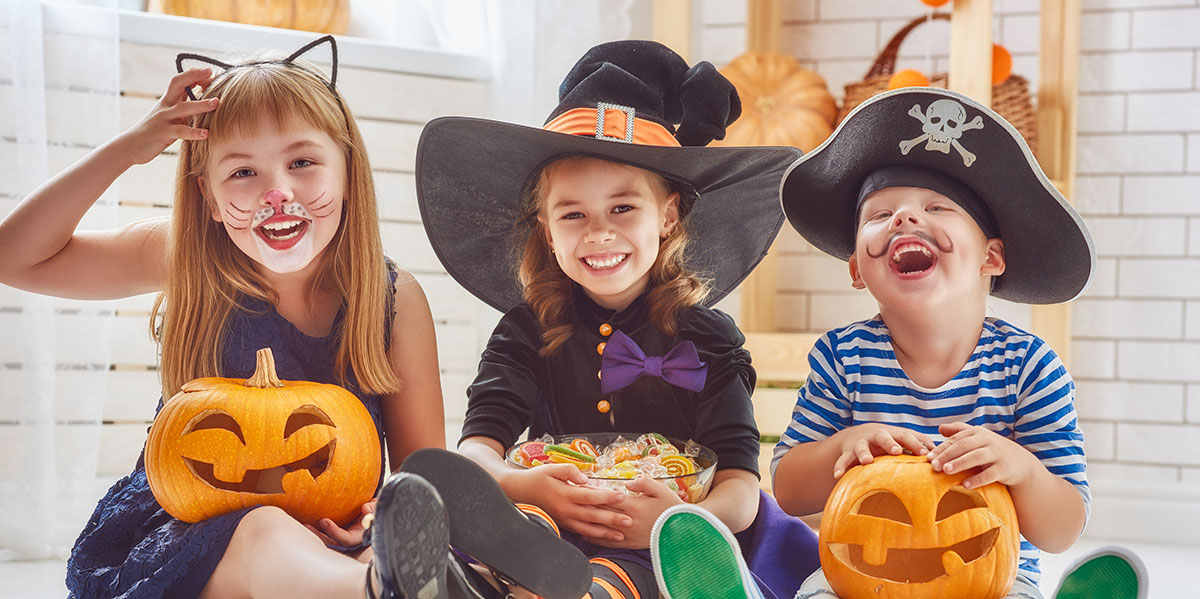 Trio of smiling children sitting together with pumpkins and candy dressed for Halloween fun as a cat, witch, and pirate