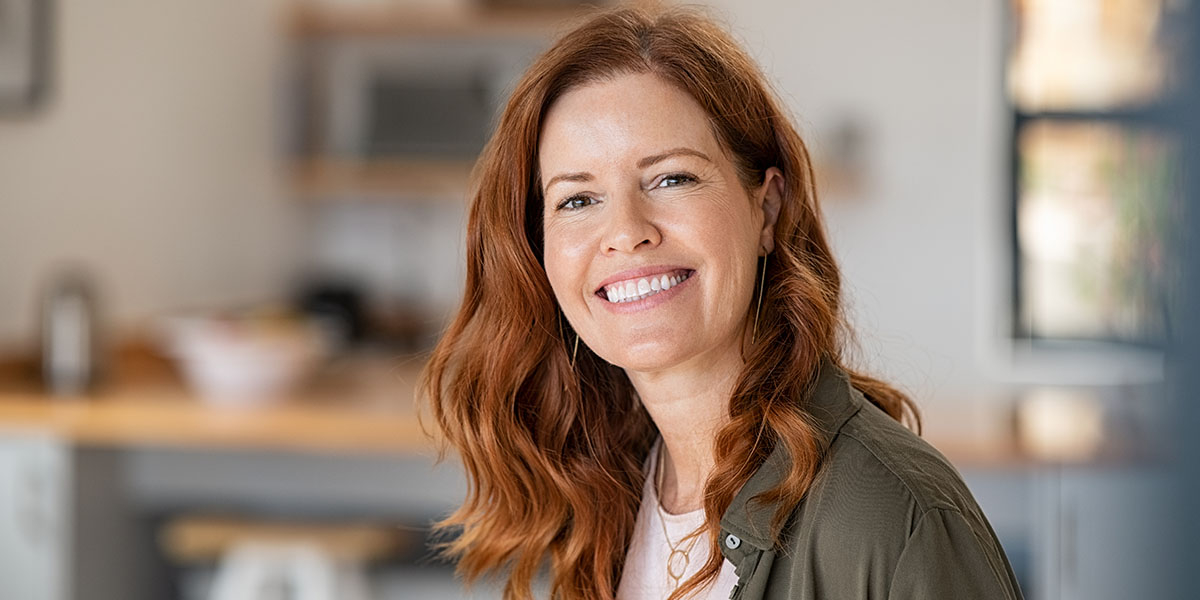 A middle-aged, red-headed woman standing in her kitchen, smiling warmly at the camera.