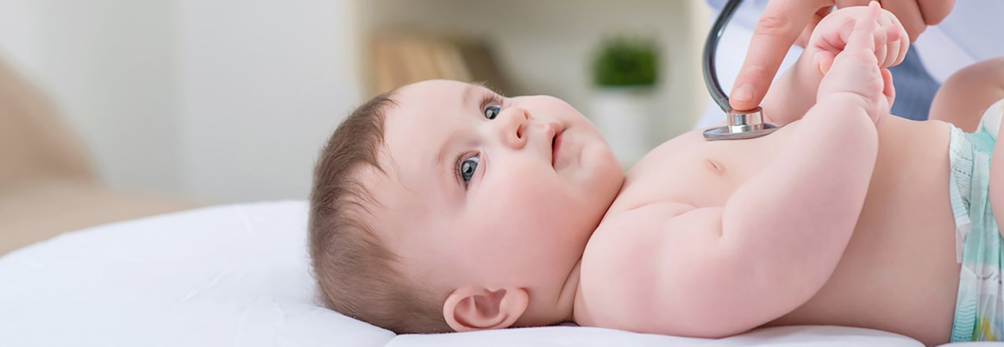 A baby lies on an exam table staring curiously upwards. A provider’s hand holds a stethoscope to the baby’s chest to check their vitals.